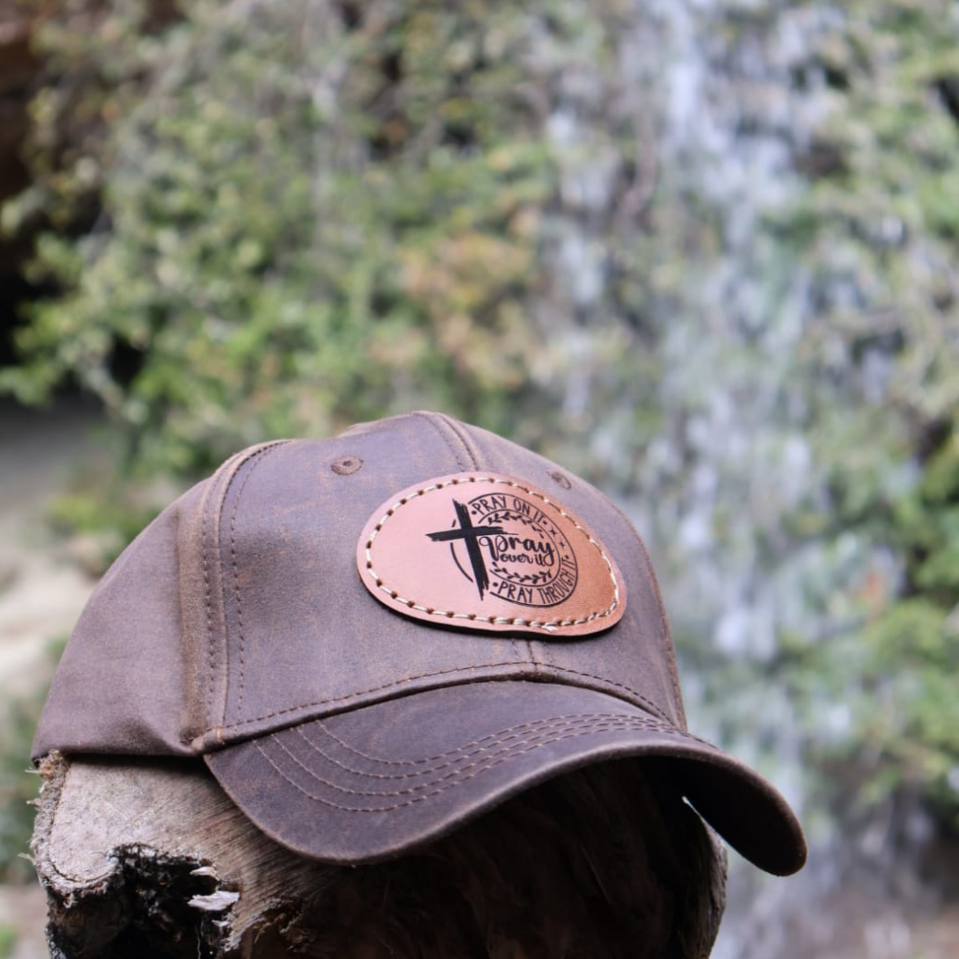 Brown cap with leather patch featuring a cross and text, placed on dried wood against a natural background.