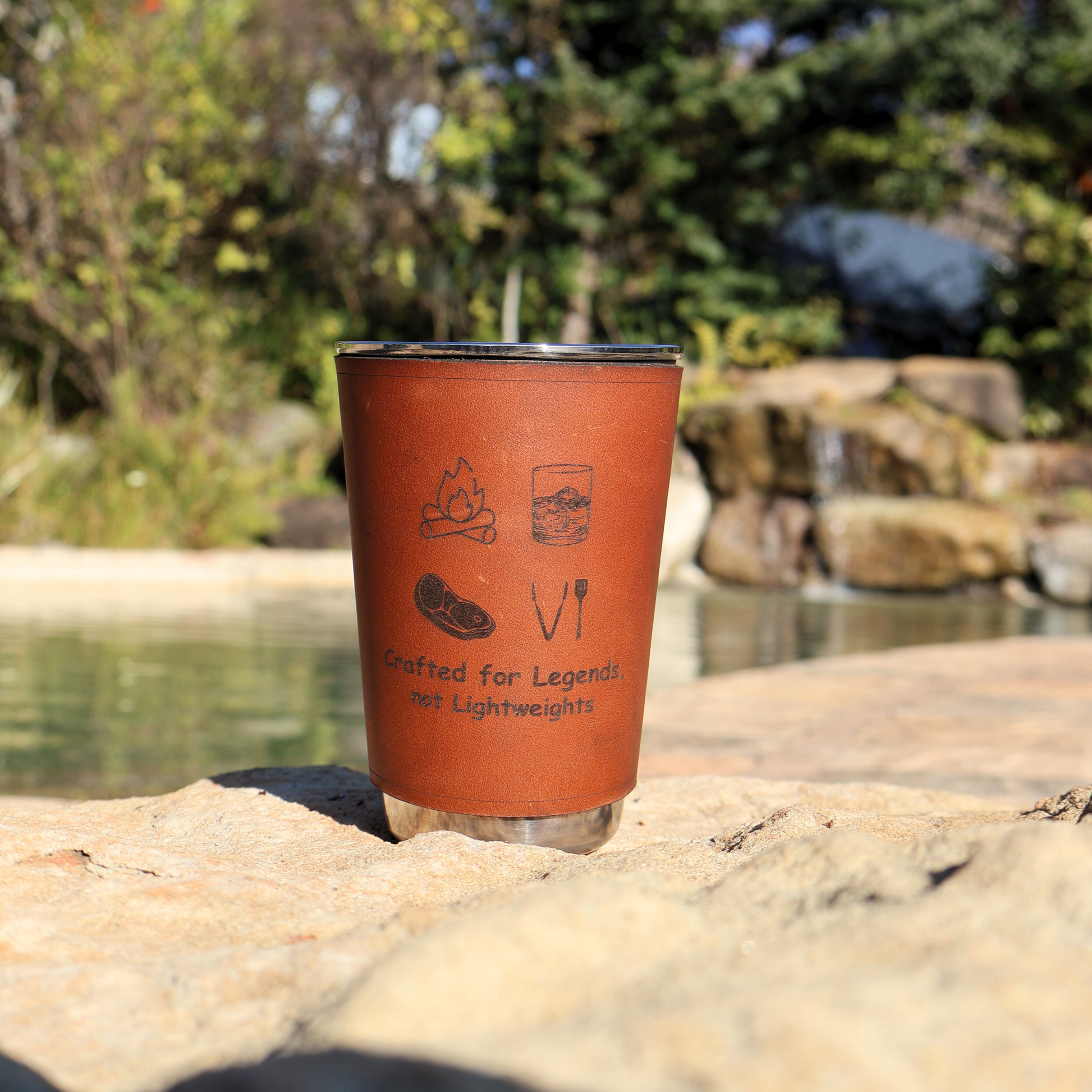 Stainless steel glass with leather sleeve engraved “Crafted for legend, not lightweights” resting on a rock near a natural pool and small waterfall