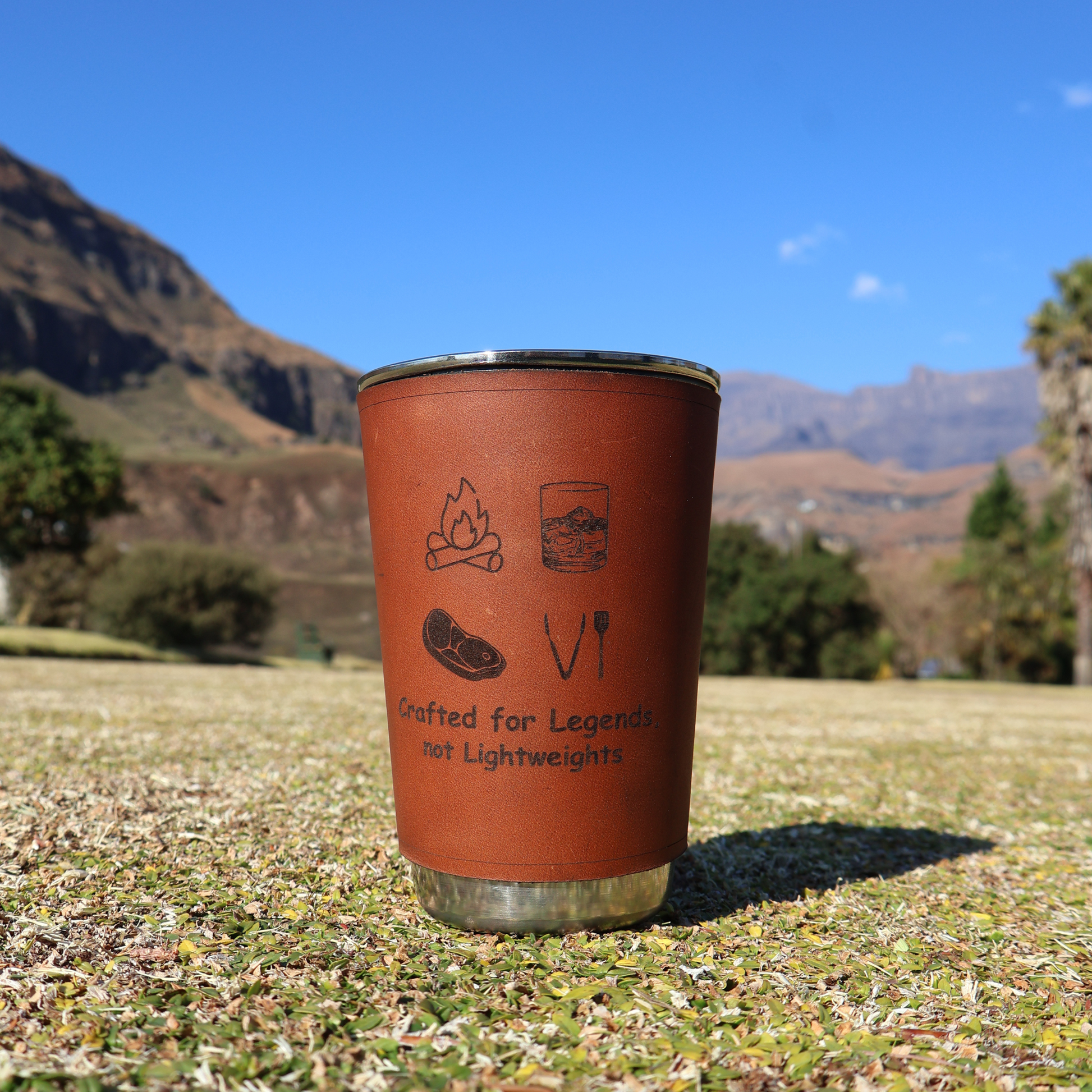 Stainless steel glass with leather sleeve engraved “Crafted for legend, not lightweights” placed on flat grass with mountains in the background