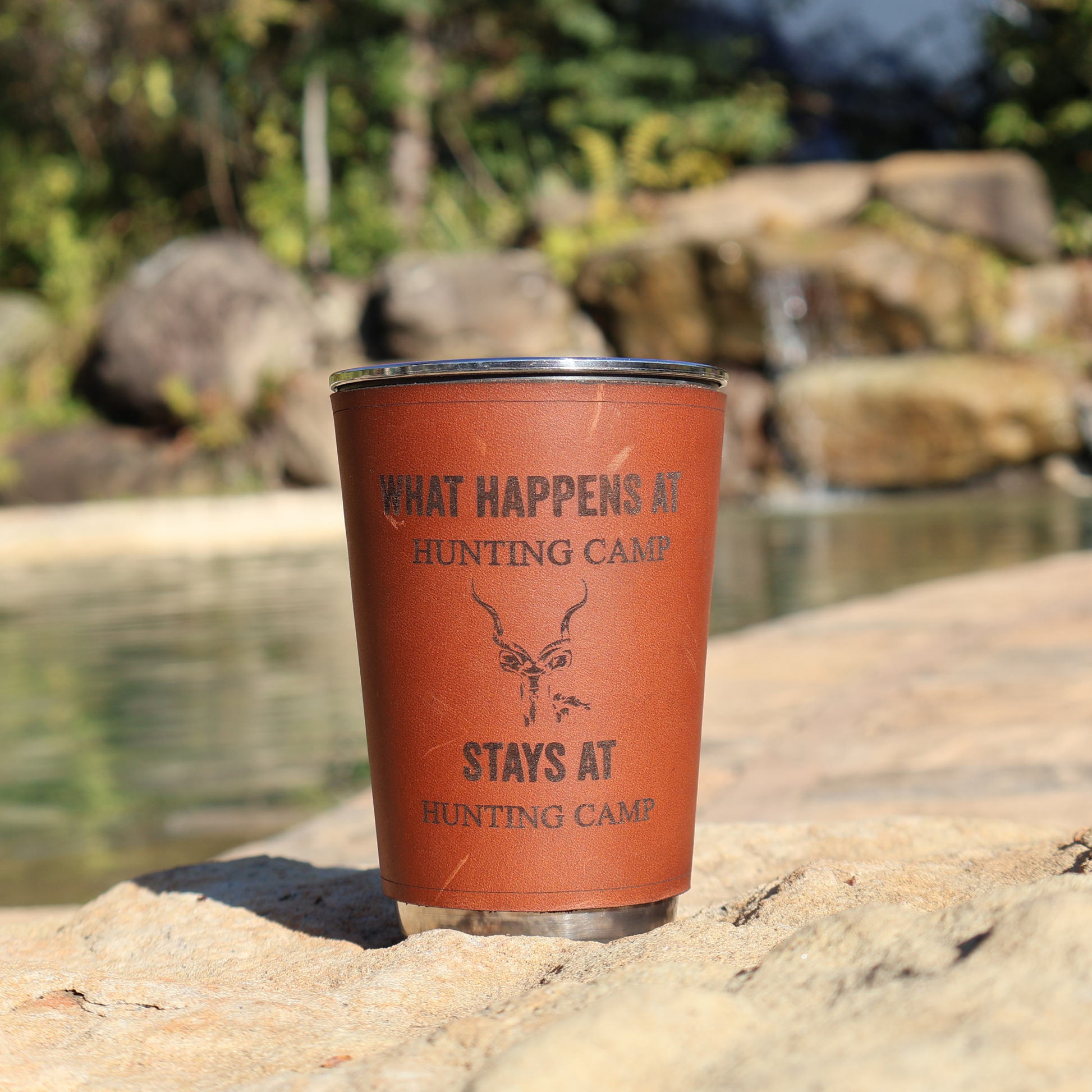 400ml stainless steel glass with leather sleeve engraved “What Happens at Hunting Camp Stays at Hunting Camp” resting on a rock near a natural pool and waterfall