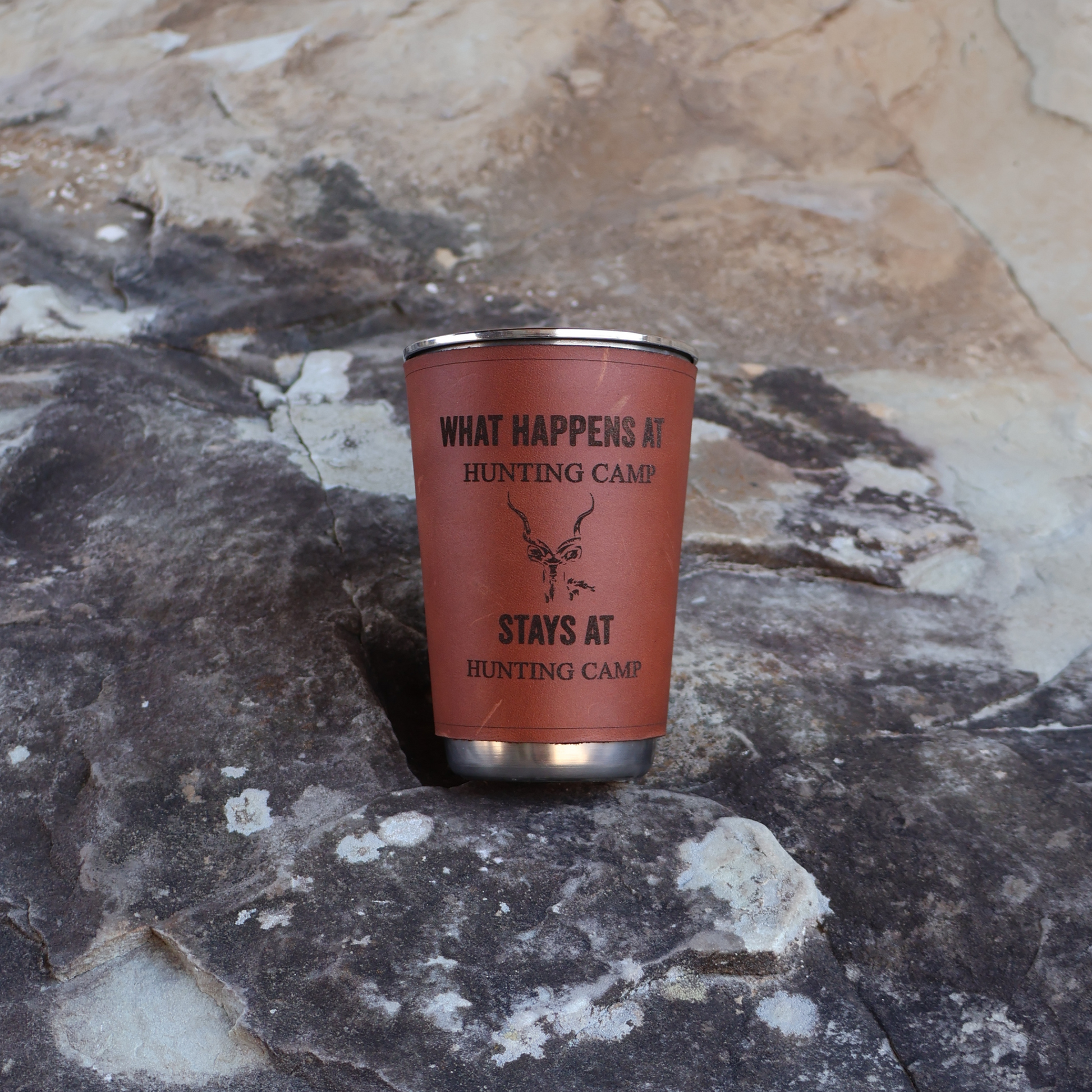 400ml stainless steel glass with leather sleeve engraved “What Happens at Hunting Camp Stays at Hunting Camp” placed on a rock with a rocky background.
