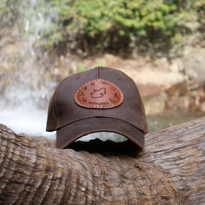 Brown oilskin cap with South Africa leather patch resting on a log in front of a waterfall, showcasing its outdoor aesthetic