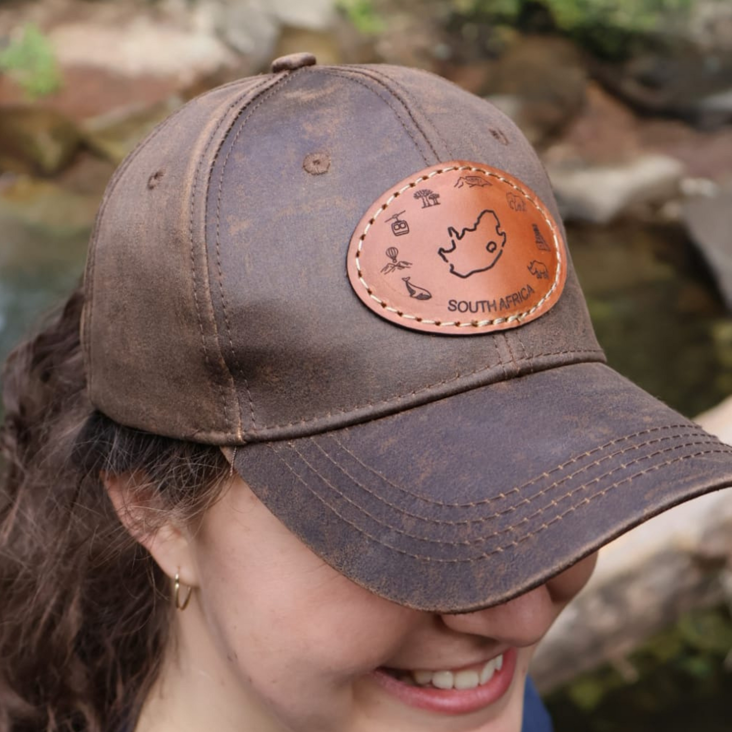 Woman with curly brown hair smiling while wearing a brown oilskin cap with South Africa leather patch