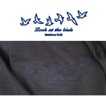 A split-screen comparison showing the clean digital blue vector graphic of flying birds and "Look at the birds Matthew 6:26" on top, paired with a close-up photo of the actual premium embroidered stitching on the navy fabric below.
