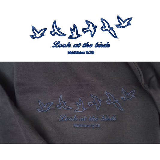 A split-screen comparison showing the clean digital blue vector graphic of flying birds and "Look at the birds Matthew 6:26" on top, paired with a close-up photo of the actual premium embroidered stitching on the navy fabric below.