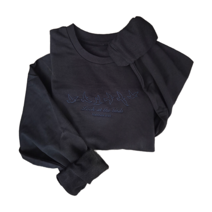 A styled flat lay of a dark charcoal grey crewneck sweatshirt featuring minimalist dark blue embroidery of six flying birds and the script text "Look at the birds Matthew 6:26" against a clean white background.