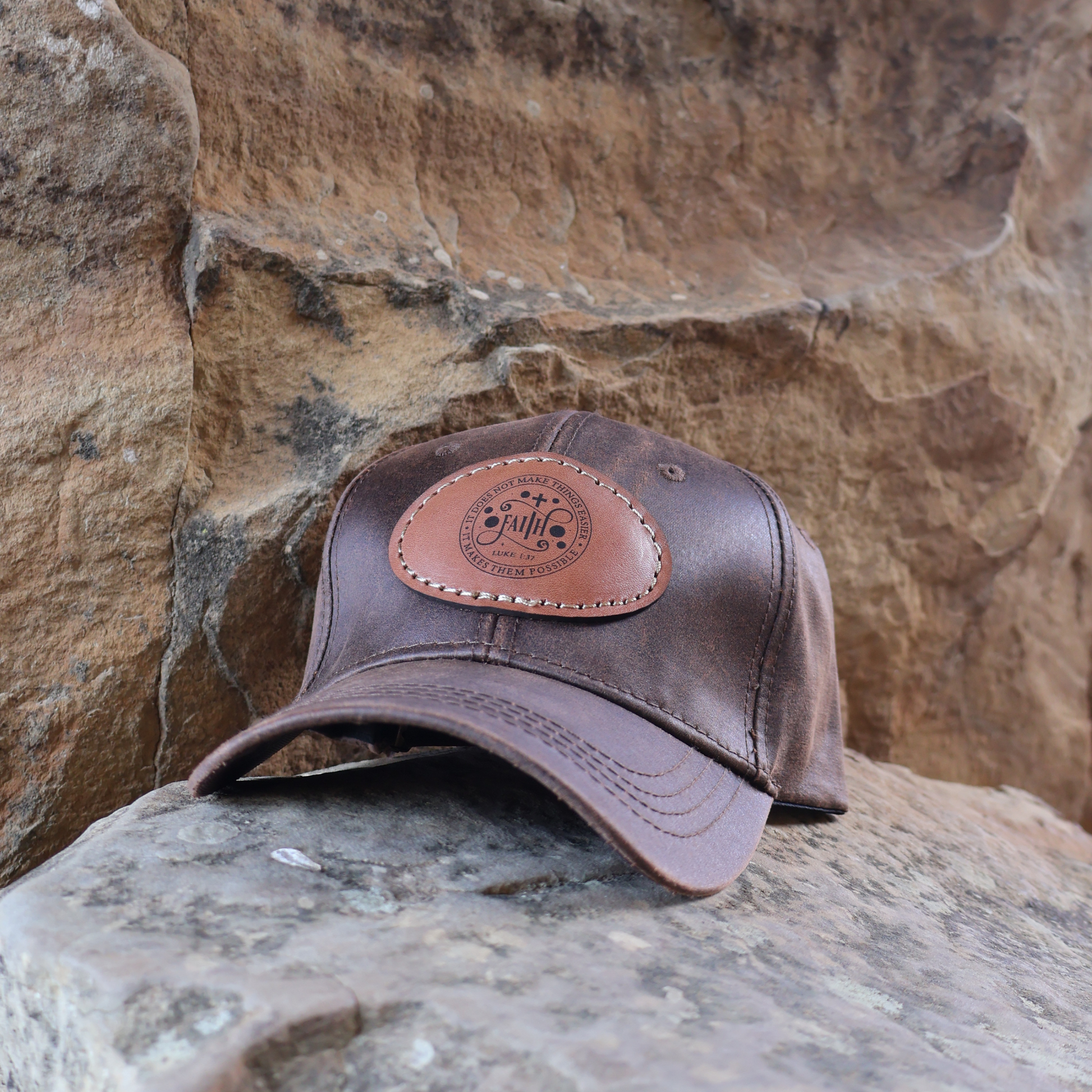 Brown oilskin cap with Luke 1:37 leather patch resting on a rock with a natural stone background.