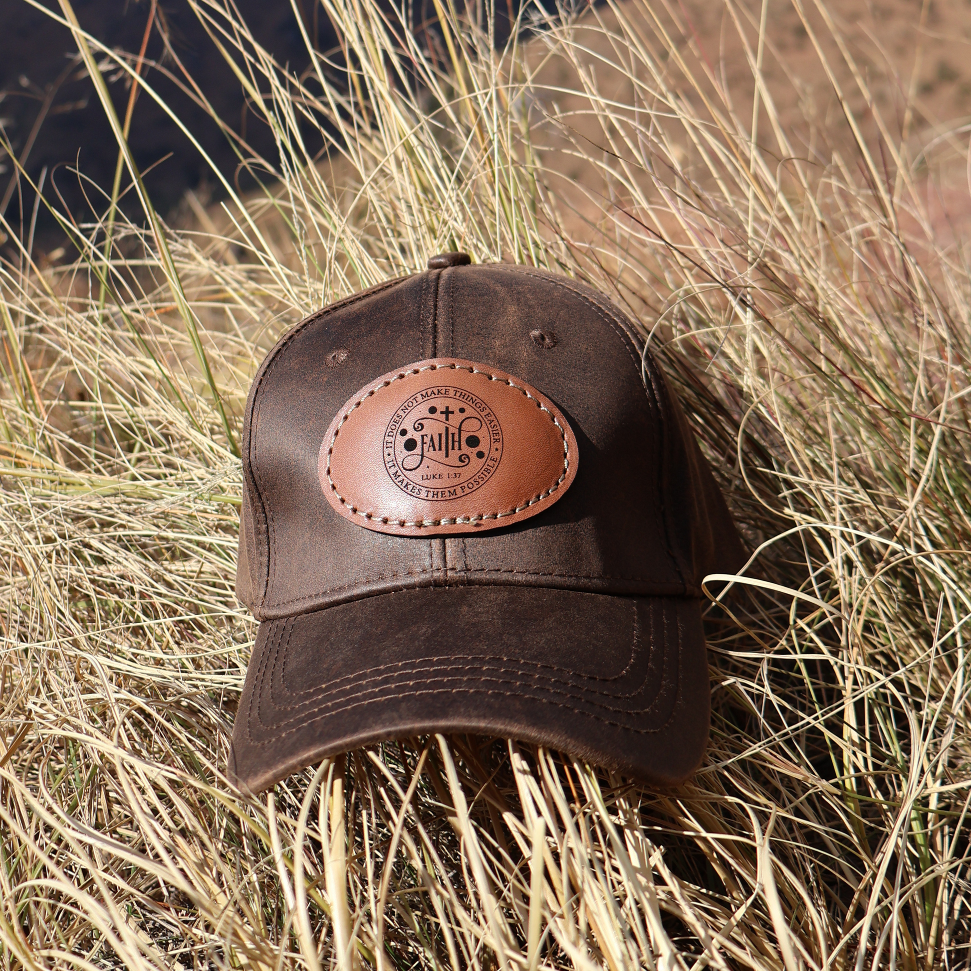 Brown oilskin cap with Luke 1:37 leather patch placed on a patch of long grass, highlighting its rugged outdoor style.