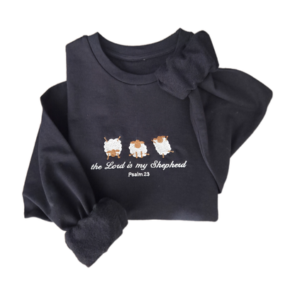 Flat lay product shot of a navy blue embroidered sweatshirt featuring three white sheep and cursive text reading "the Lord is my Shepherd Psalm 23". The cozy christian jumper is artistically folded against a clean white background.