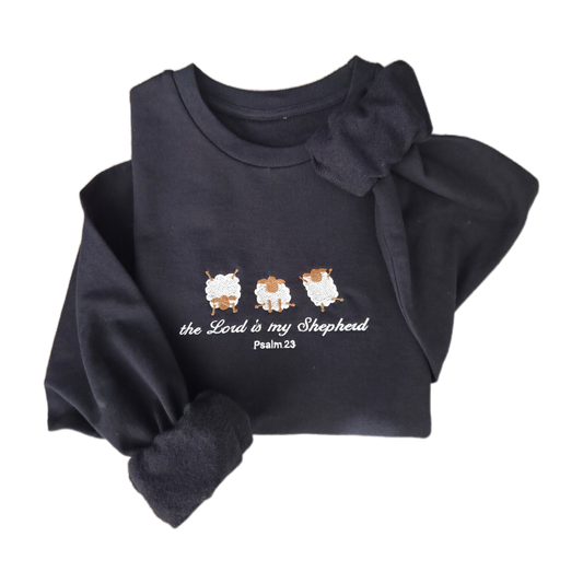 Flat lay product shot of a navy blue embroidered sweatshirt featuring three white sheep and cursive text reading "the Lord is my Shepherd Psalm 23". The cozy christian jumper is artistically folded against a clean white background.